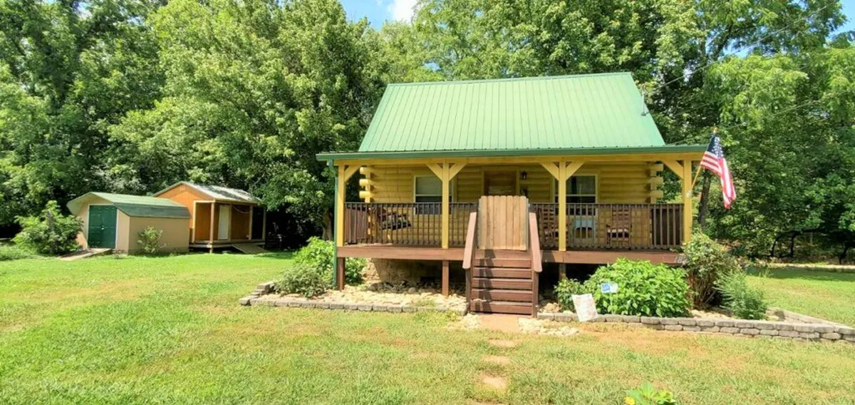 The yellow house with a green roof and a welcoming front porch is the centerpiece, with a visible storage shed and garden space. The porch is set up for relaxing and socializing, evoking a sense of friendly, homely atmosphere in the outdoors.
