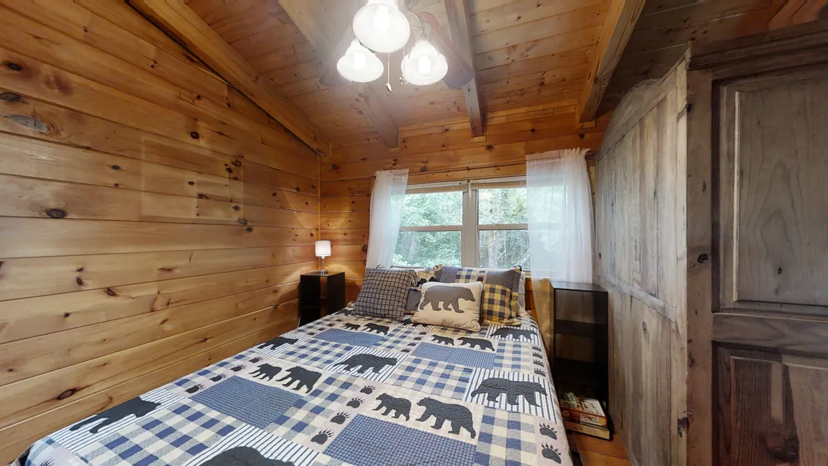 A tranquil bedroom scene with a comfortable bed facing a window, wooden walls, and a soft lighting fixture. The gentle sunlight suggests a peaceful retreat, ideal for resting after a day spent outdoors.