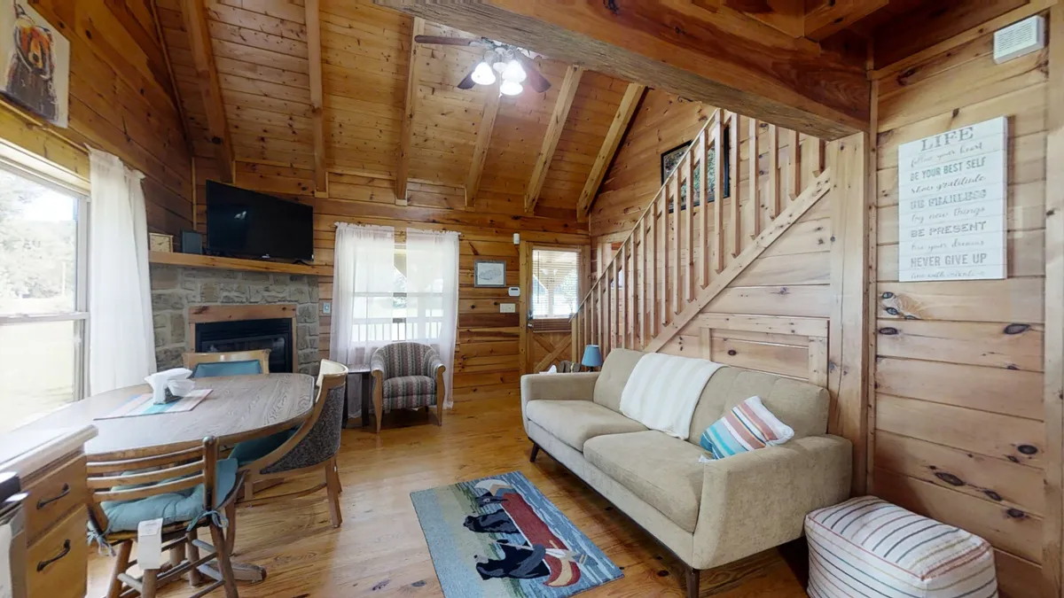 A cozy interior scene centered around exposed wooden beams, a striking stone fireplace, a comfortable sofa, and a dining table bathed in daylight. The space exudes warmth and relaxation, ideal for unwinding after a day of adventure or enjoying time together inside the cabin.