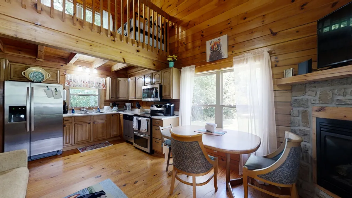 This image displays a warm and inviting open-plan space that seamlessly connects the kitchen, dining area, and living room. You can see the kitchen island in the foreground, a dining table set for family meals, and a cozy fireplace adjacent to large windows that flood the space with natural light. The arrangement captures both gathering and relaxation spaces, and highlights the key features of the main level.