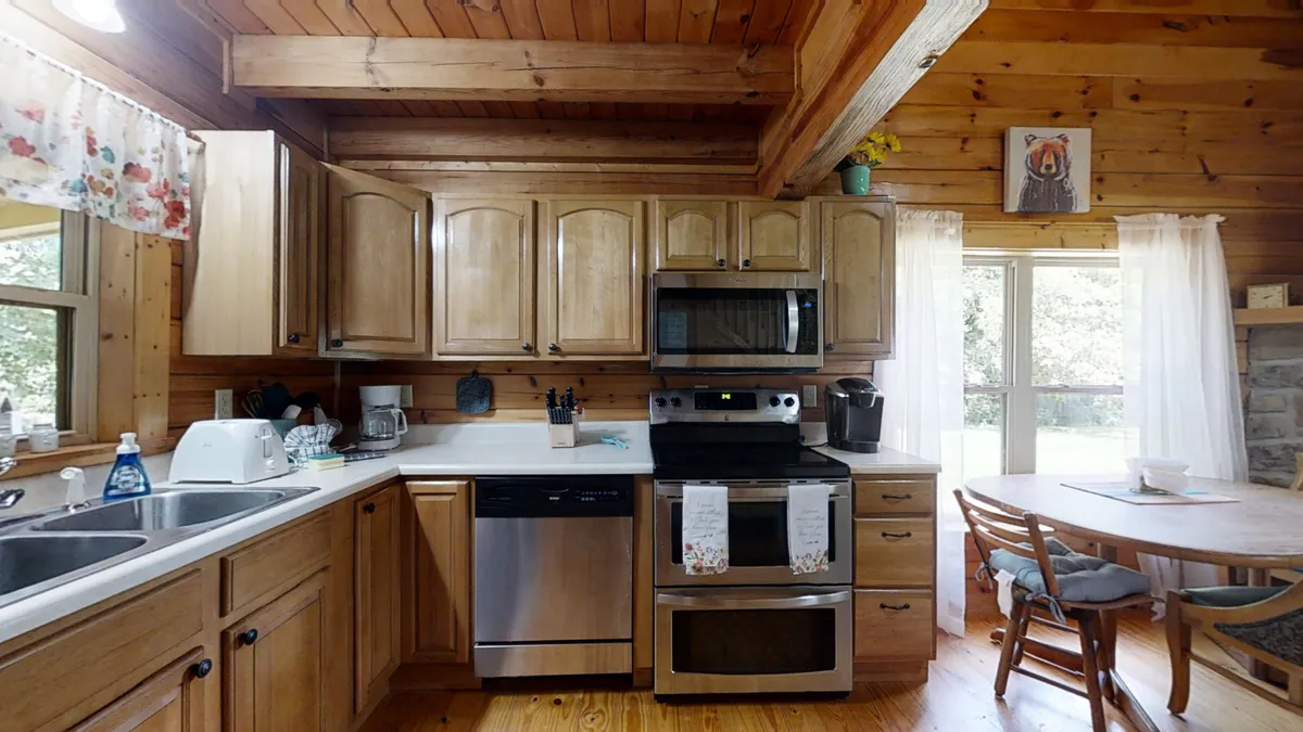 A welcoming view of the Yellow Cabin's kitchen and dining area, featuring warm wooden cabinetry, sleek stainless steel appliances, a cozy dining table, and a window draped with sheer curtains. The atmosphere is inviting and reflects the warmth of gathering indoors during cooler months.