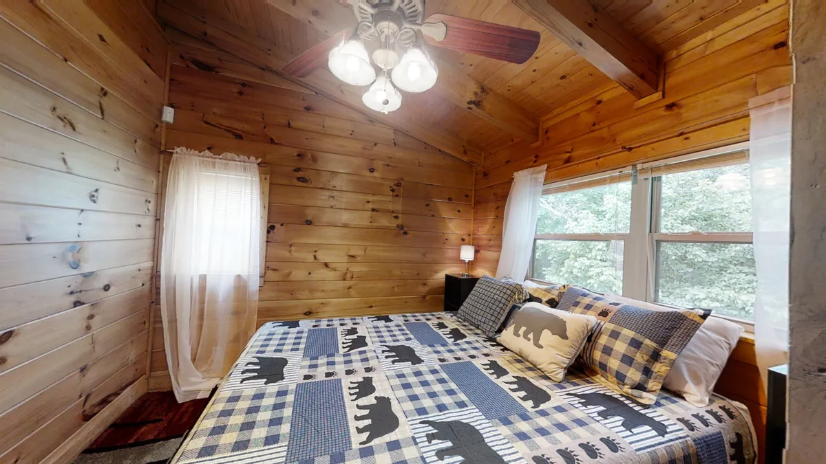 A warmly lit bedroom featuring bear-themed bedding, expansive windows allowing natural light to pour in, and rustic wooden walls and ceiling. The space feels cozy, private, and deeply connected to the cabin’s mountain surroundings.