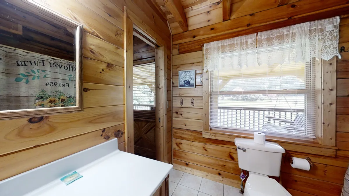A tranquil bathroom with wooden walls, a mirror, a sink beneath a window with a lace curtain, and a toilet. The clean, inviting ambiance is enhanced by natural light, making it a perfect space for refreshing after a day outdoors.