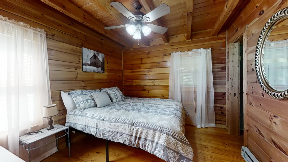 This image features a serene bedroom with wooden walls, a plush bed adorned with soft linens, a ceiling fan overhead, and a mirror reflecting warm natural light. The tranquil atmosphere is perfect for relaxation and captures the essence of a restful retreat.