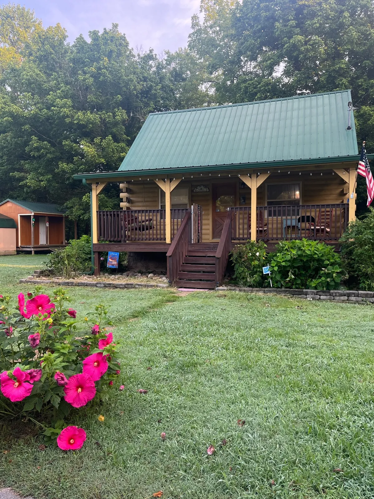 A wide wooden porch accented with bright flowers, a green metal roof, and the American flag. Comfortable seating options invite guests to relax and enjoy views of the yard and garden, with hints of the fire pit area nearby.