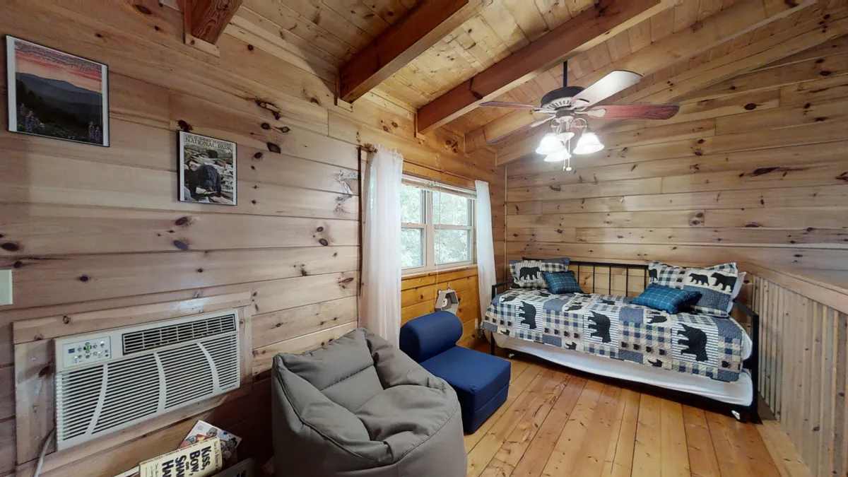 A cozy loft space featuring a bed with plaid bedding, exposed wooden beams, a bean bag chair, and a window letting in natural light. The setting is comfortable and casual, ideal for flexible sleeping arrangements and late-night conversations.