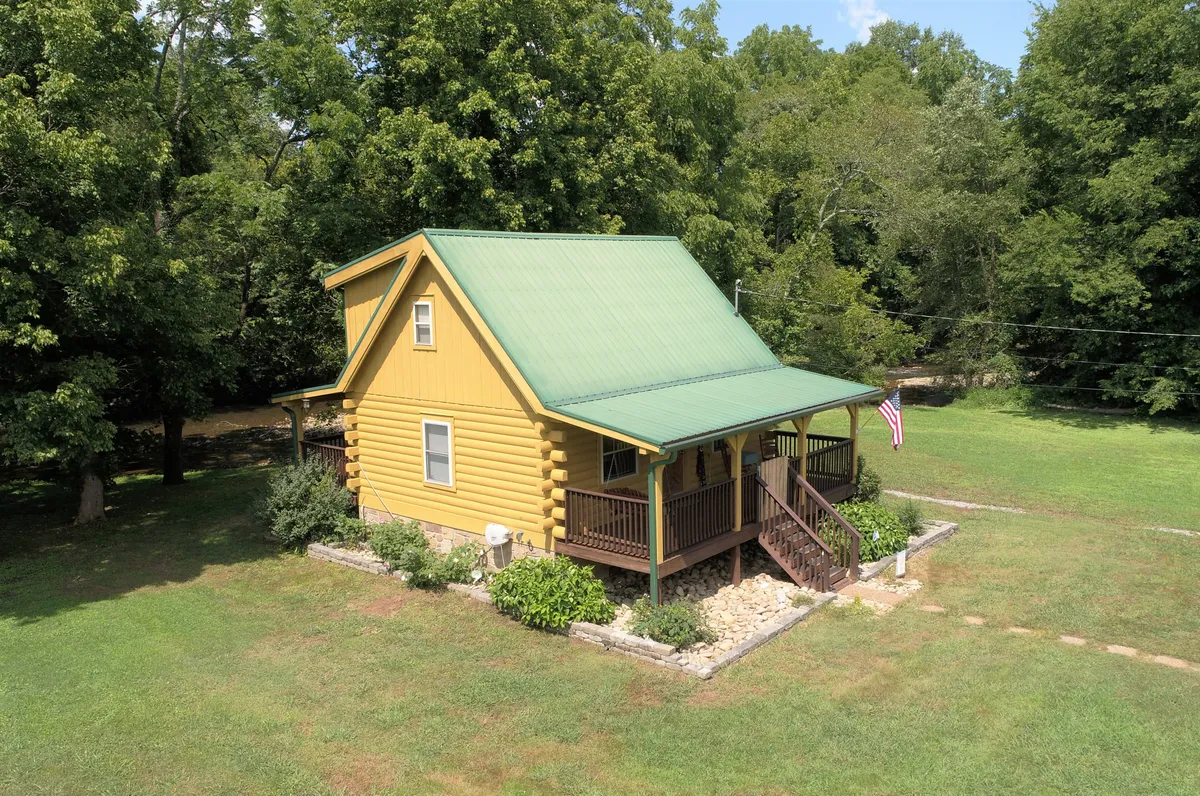 This exterior shot features the yellow log cabin with a green metal roof, a welcoming wooden staircase, and an American flag. The sunny porch suggests ease of arrival and departure, and the visible front entry underlines the self-check-in process. The paved area in front is clear and accessible, hinting at motorcycle-friendly parking.