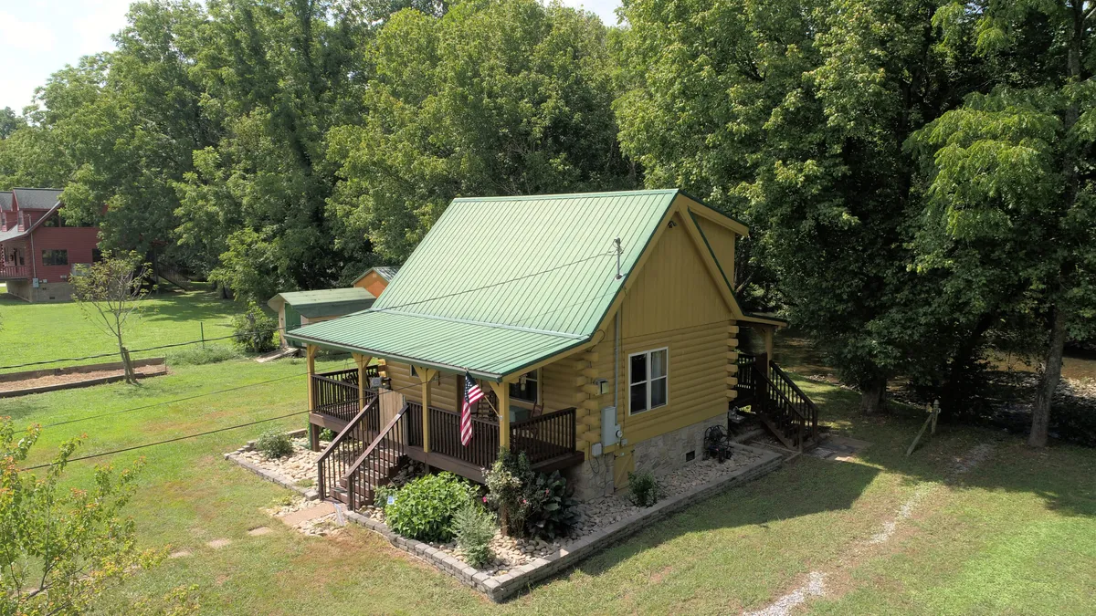 Another angle of the cabin's exterior, highlighting the landscaped garden, green metal roof, and inviting front porch with steps. The setting is tranquil and accessible, reinforcing the ease of arrival and the option to relax outdoors with pets or family.