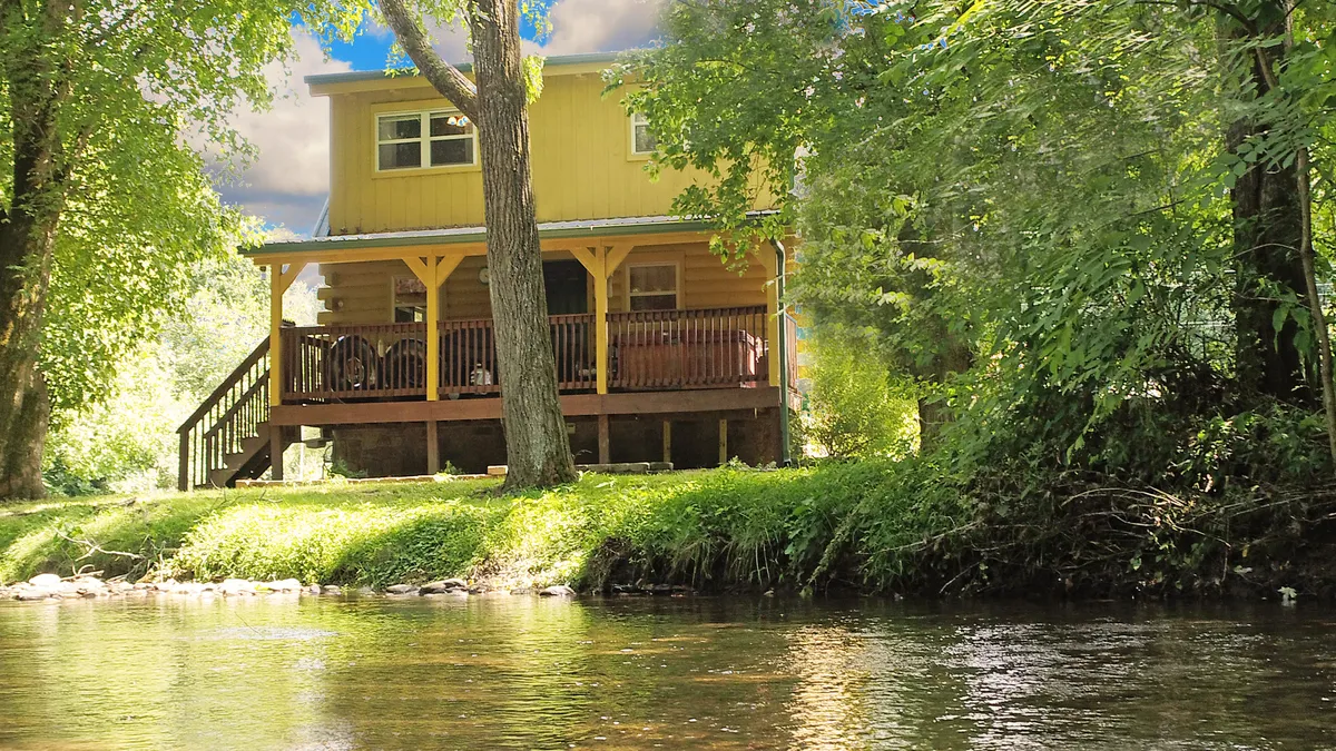 The image shows a charming yellow cabin perched beside a tranquil river, surrounded by lush trees. A spacious porch overlooks the water, inviting guests to relax while taking in serene sunset views. The natural landscape hints at opportunities for fishing, wildlife watching, and enjoying peaceful moments in nature.