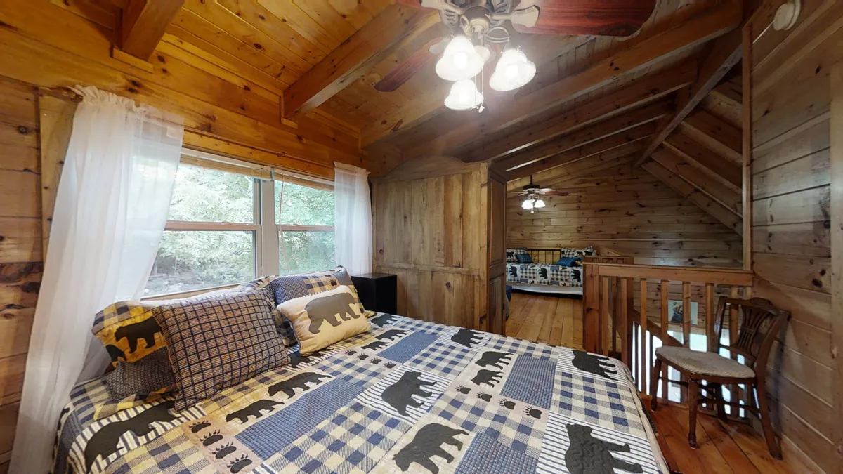 The image highlights a cozy bedroom with a bear-patterned blanket on the bed, large windows letting in ample natural light, and rustic wooden beams and paneling. The windows provide a view to the outdoors, suggesting a seamless connection between indoor comfort and nature just outside.