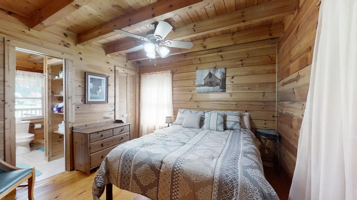 A welcoming bedroom with a patterned bedspread, wooden walls, ceiling fan, and artistic touches on the wall. Sunlight filters through the windows, creating an inviting and peaceful space perfect for winding down after a day of adventure.